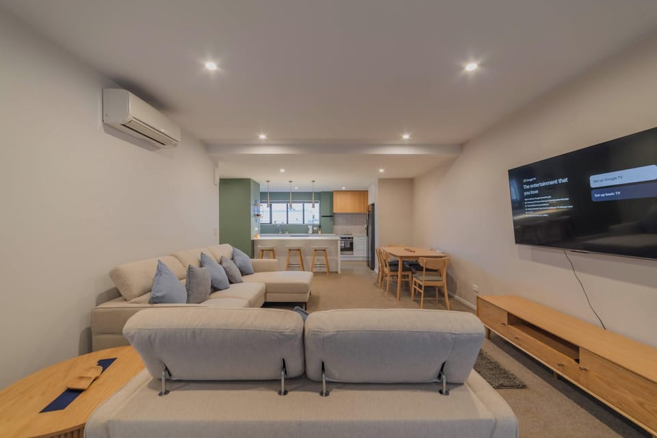 Bright open-plan living area with generous sofa seating, wall-mounted TV, media console, dining table and full kitchen beyond.