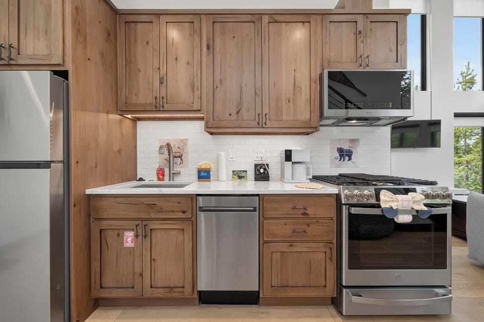 Your fully equipped kitchen invites culinary adventures with its warm wood cabinetry, modern stainless steel appliances, and bright natural light streaming through large windows overlooking the greenery outside.