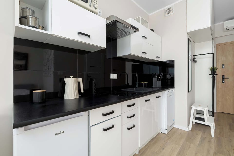 A compact kitchenette displays glossy white lower cabinets, black upper units, and a dark splashback. The wooden flooring extends past the front door.