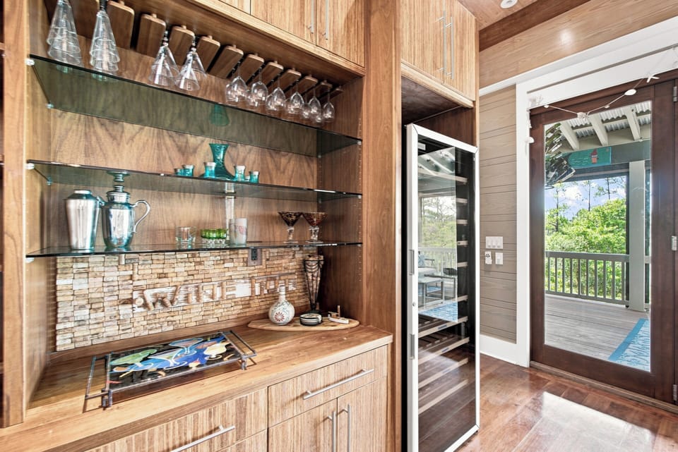 Your elegant bar area features glass shelving to display glassware and cocktail essentials, with a stunning natural backdrop visible through glass doors leading to the deck.