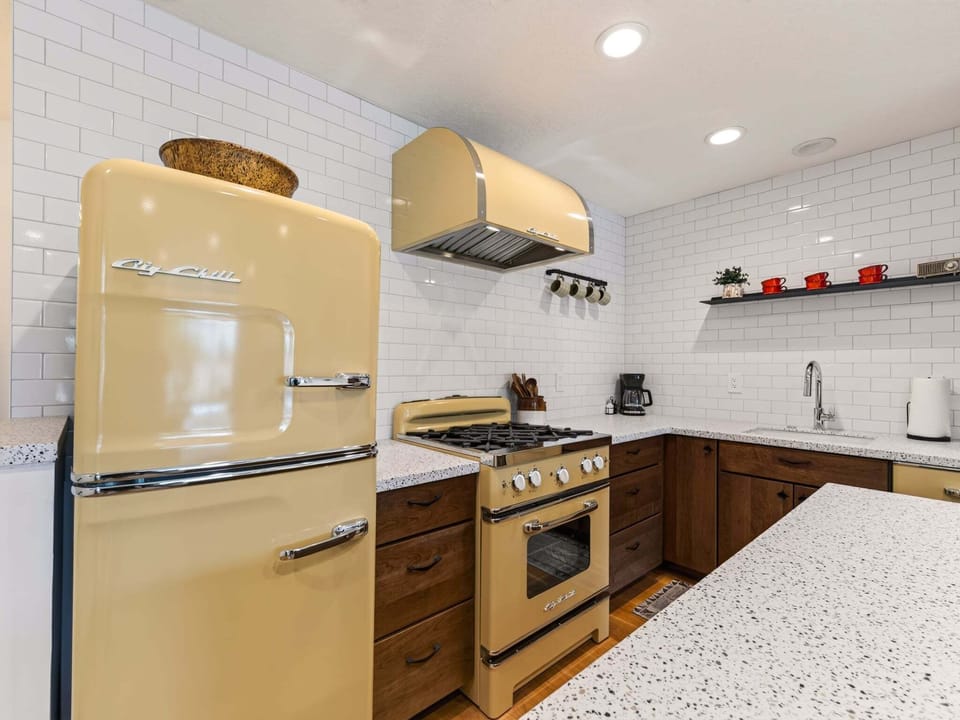 A retro inspired kitchen with modern convenience, featuring warm wood cabinetry, vintage style appliances, quartz countertops, and open shelving. Fully stocked with cookware, utensils, and everything you need to prepare meals with ease.