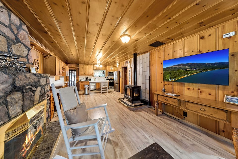 Warm living space wrapped in rich floor-to-ceiling knotty pine, complete with a wall-mounted TV, white rocking chair, and classic wood-burning stove.