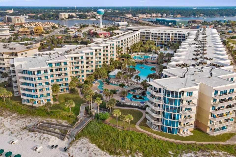 Front drone view of Waterscape Resort featuring the full beachfront property, resort pools, lazy river, tropical landscaping, and the bridge leading to Fort Walton Beach on beautiful Okaloosa Island along Florida’s Emerald Coast.