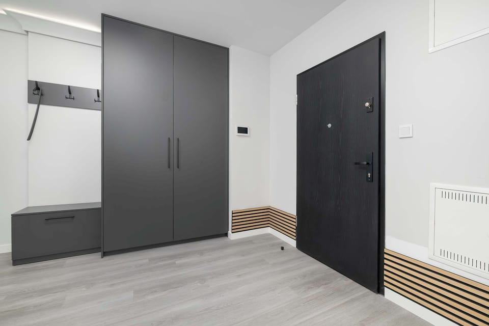 A dark entrance door and a second interior door visible in a simple hallway with light wood flooring. The minimalist space provides access to the apartment's rooms.