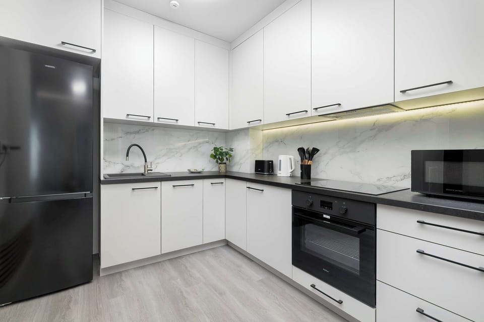 A compact modern kitchen fitted with white cabinetry, a dark refrigerator, and built-in cooking appliances. The minimalist design provides a clean and functional cooking space.