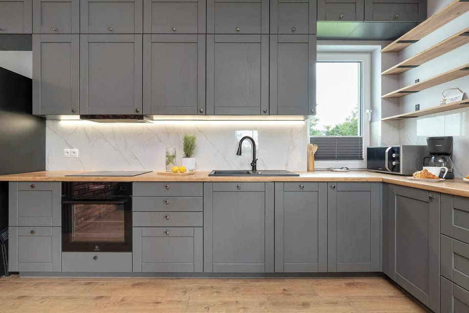 The kitchenette workspace features sleek matte-grey cabinets, an integrated electric oven, and an induction cooktop. Neutral worktops and open wooden corner shelving offer plenty of prep room. A window above the counter brings in fresh daylight.