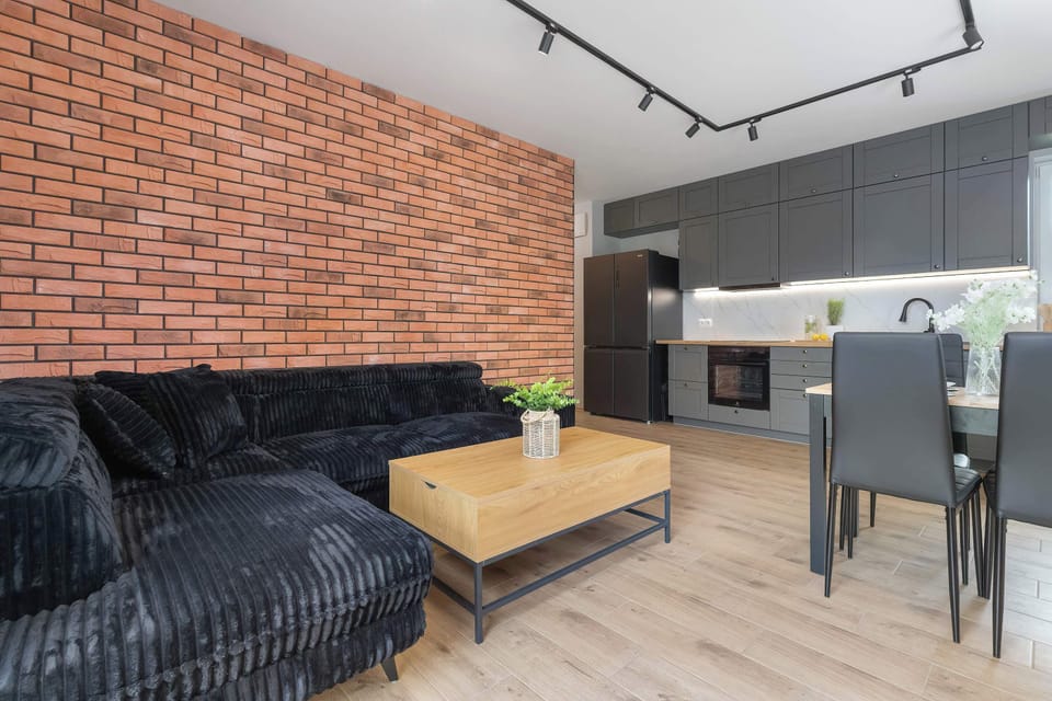 A wide-angle perspective connects the lounge area with the adjoining dining space and kitchenette. Black ceiling track spotlights provide sleek, directional illumination. The brick accent wall anchors the room's warm modern design.