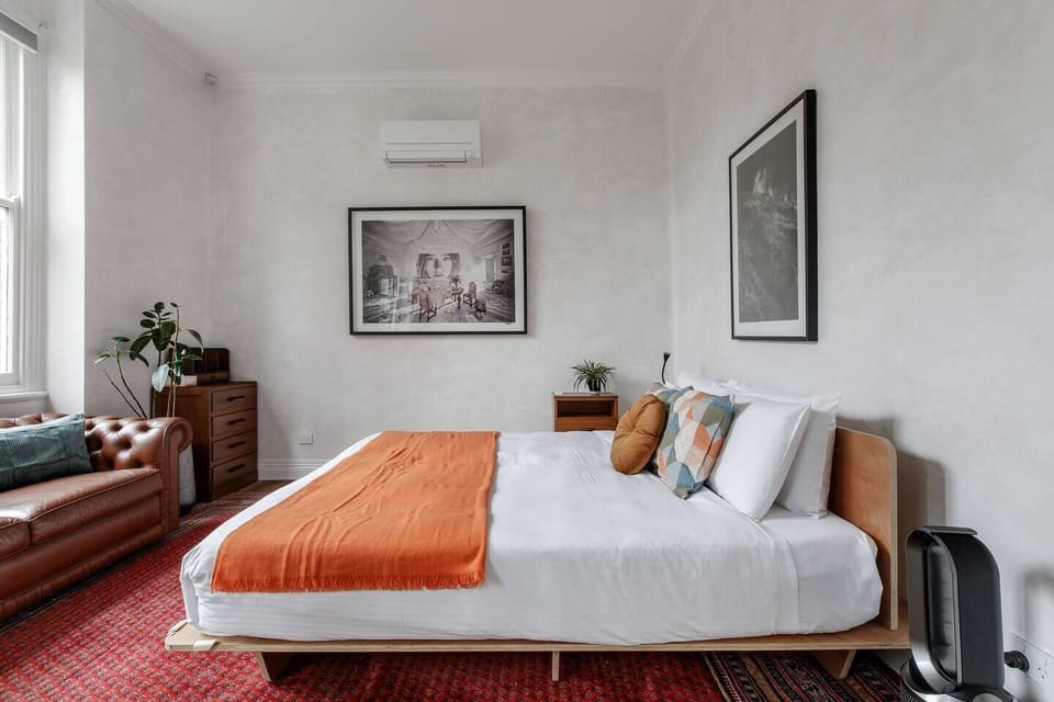 The primary bedroom offers a king bed, wall art, a leather Chesterfield sofa, clothing storage, and split-system air conditioning for ultimate comfort.