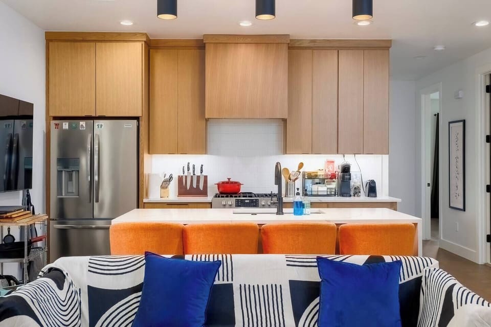 This modern open-concept space seamlessly blends living room and kitchen areas. Features include a gray sectional sofa, wall-mounted TV, unique zigzag bookshelf, and honey-toned wooden cabinetry with white countertops and stainless appliances.