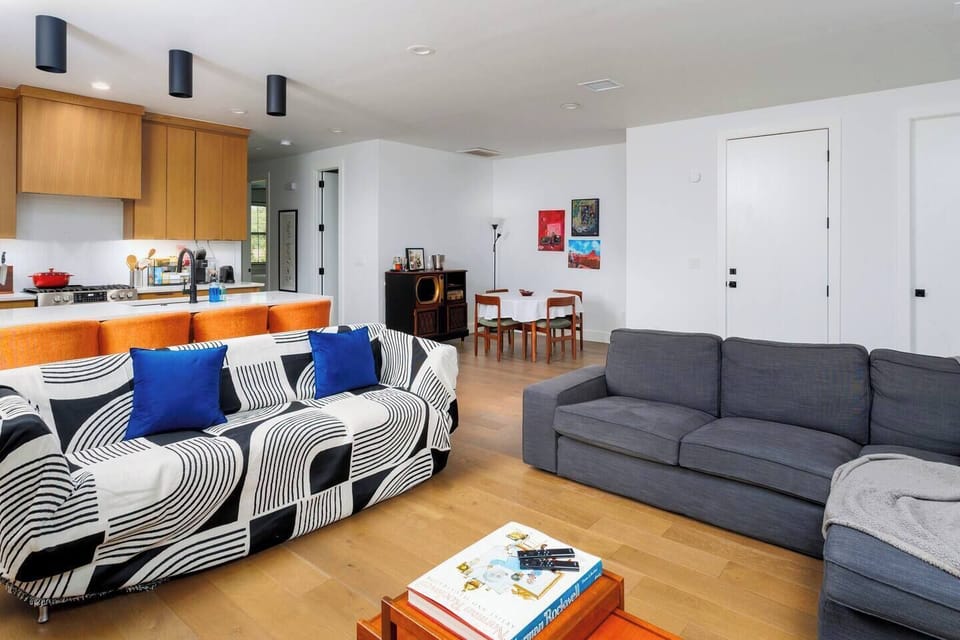 This modern open-concept space seamlessly blends living room and kitchen areas. Features include a gray sectional sofa, wall-mounted TV, unique zigzag bookshelf, and honey-toned wooden cabinetry with white countertops and stainless appliances.