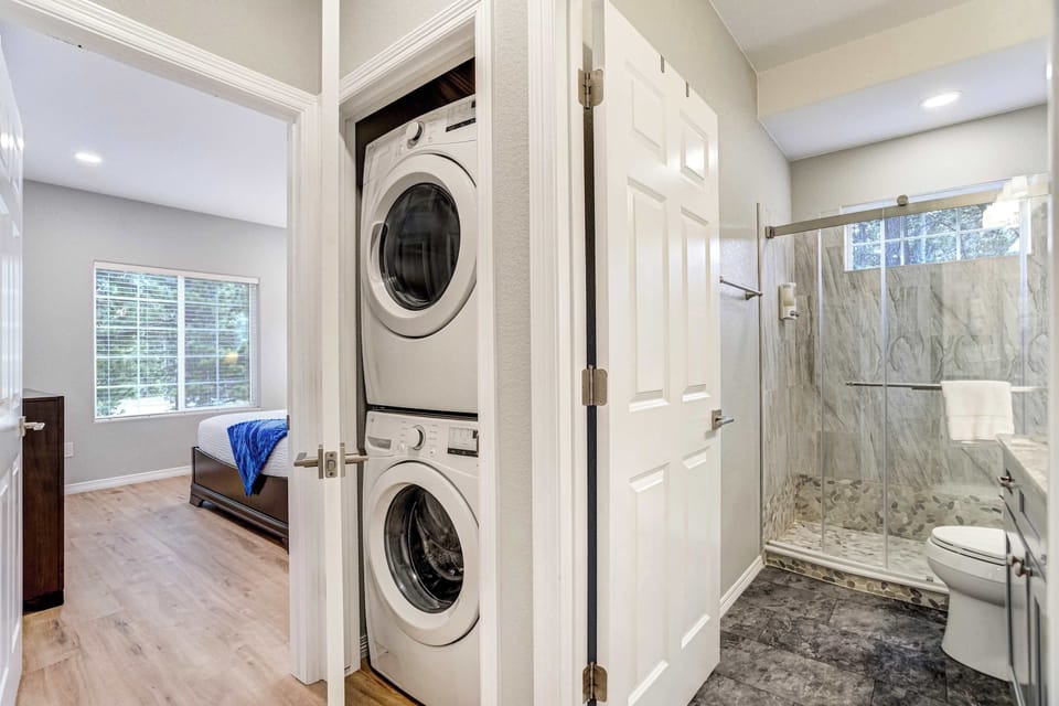 A stacked washer and dryer tucks into the hallway just outside the bedroom, so packing light is easy and hiking clothes never pile up. Just beyond it, the full bathroom opens with a glass enclosed walk-in shower and warm light.
