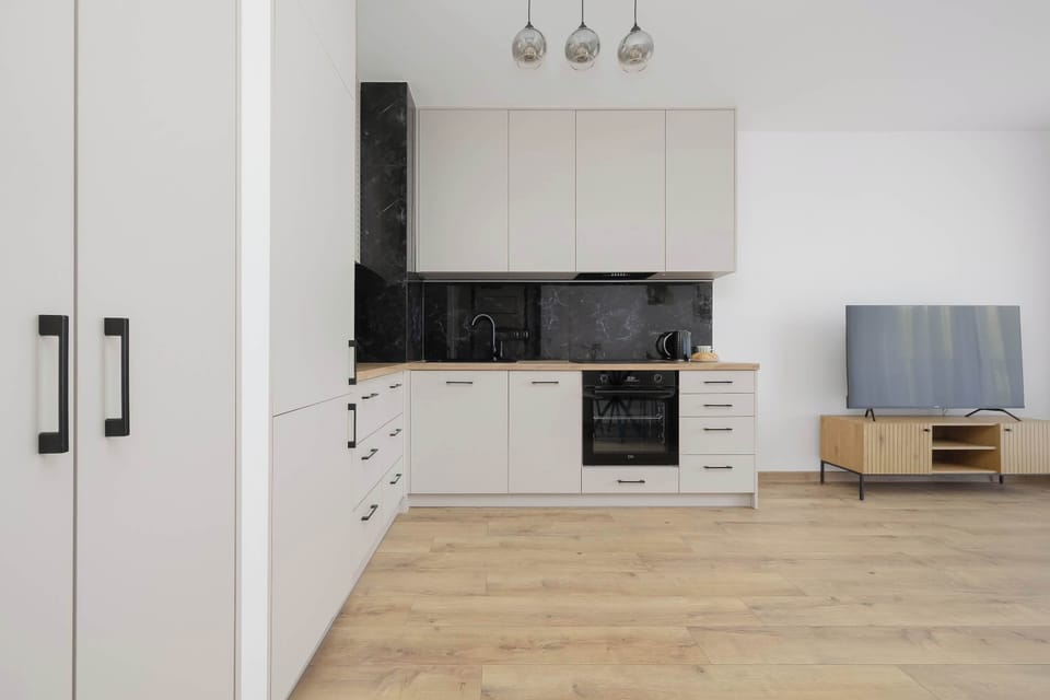 Modern kitchenette with white cabinets, a built-in oven, a black countertop and backsplash, a refrigerator and a wooden floor.