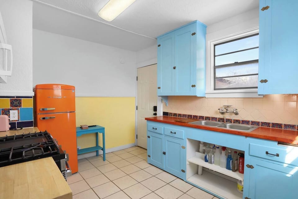 Bright, retro-inspired kitchen with bold blue cabinetry and a statement orange fridge. Enjoy a sunny space with full cooking amenities, tiled backsplash, and plenty of counter space—perfect for slow mornings and home-cooked meals.