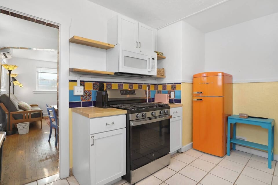Charming vintage kitchen with a bold orange fridge, gas stove, and colorful tiled backsplash. Fully equipped for easy cooking, with open shelving and a bright, airy layout that flows into the living space.