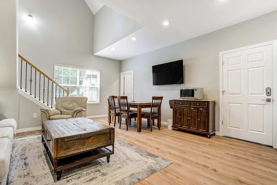 The main level opens up with living, dining, and the kitchenette all sharing one bright space. A wall mounted Smart TV faces the sectional, and light hardwood runs underfoot beneath a rustic coffee table set on iron casters.