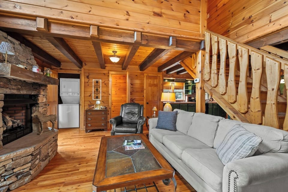 A vaulted‑ceiling living room with a stone fireplace, oversized windows, and a large smart TV—your cozy retreat for movie nights and morning coffee with a view.