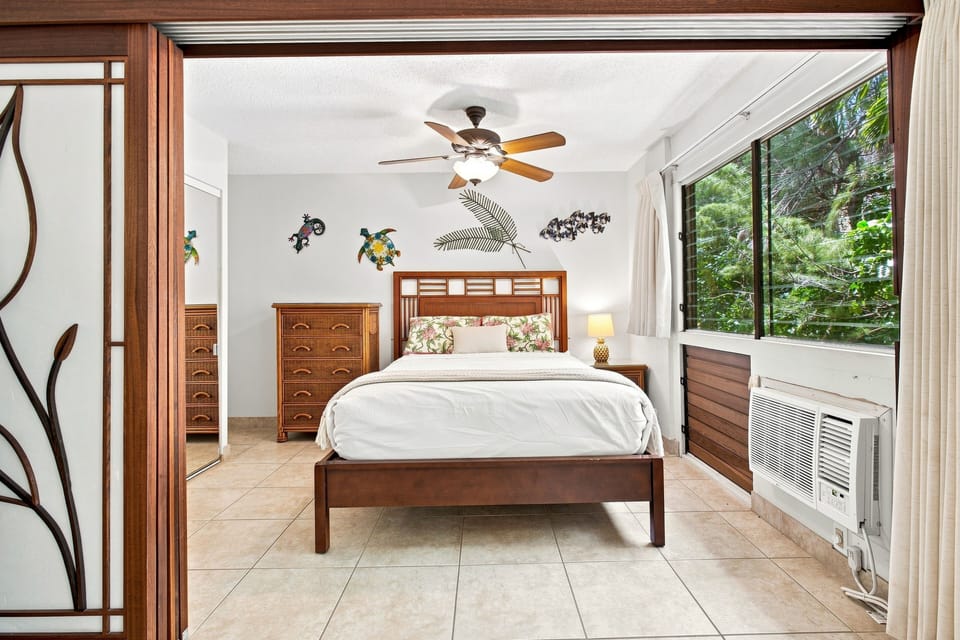 Drift off in your inviting bedroom with warm tropical touches, soothing ceiling fan, and lush garden views through floor-to-ceiling windows—your peaceful Hawaiian retreat awaits.