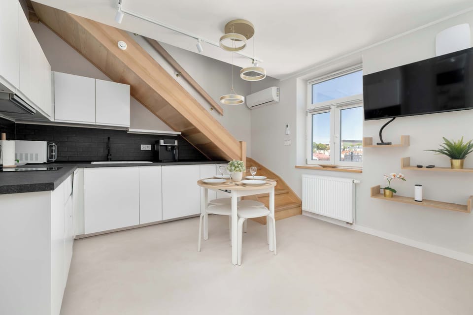 A functional kitchen and dining space opens up under angled ceiling beams, equipped with white cabinetry, a dining table, and a wall-mounted TV. The bright walls and window keep the space airy and inviting.