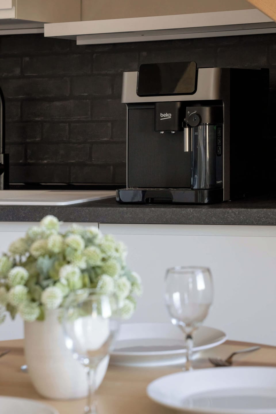 A close-up view highlights a modern coffee machine set on the dark kitchen countertop alongside fresh flowers in a vase and glassware. These thoughtful details provide a welcoming touch for morning coffee.