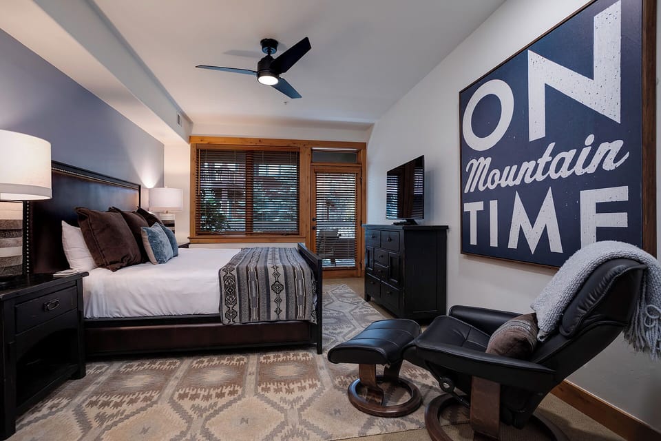 Unwind in your cozy mountain retreat with a plush bed, soft lighting, and a comfy chair. Drift off under the ceiling fan as natural light filters through wood-framed windows.
