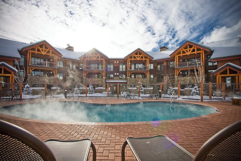 This alpine-style property features a heated outdoor pool with wooden deck, surrounded by mountain lodge buildings with exposed timber beams and balconies. The winter setting and architecture create an inviting ski resort atmosphere.