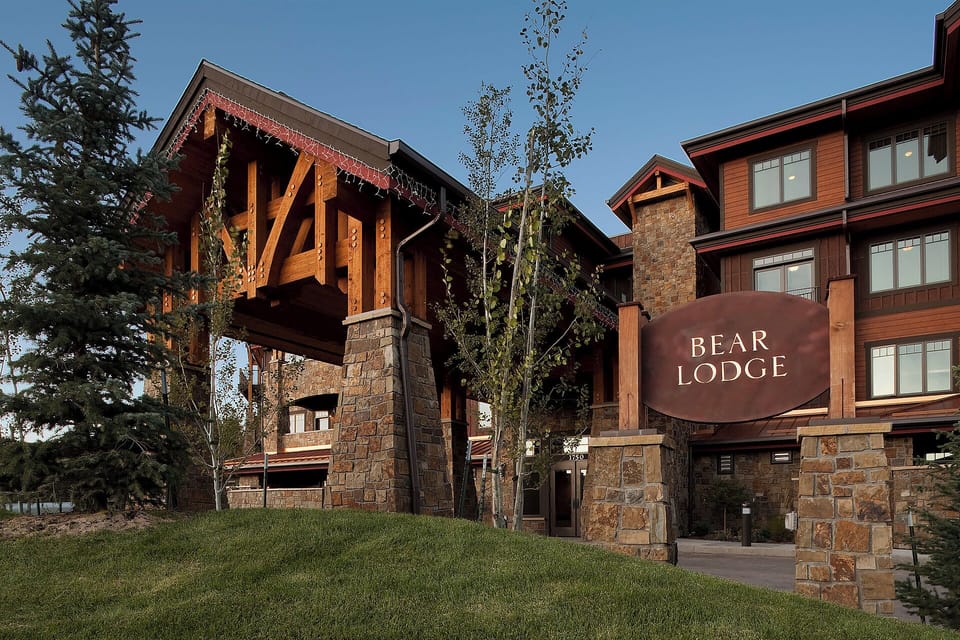 The Bear Lodge stands as a striking multi-level mountain retreat with hand-carved timber details, stone masonry, and warm wood tones nestled among tall pines and aspens.