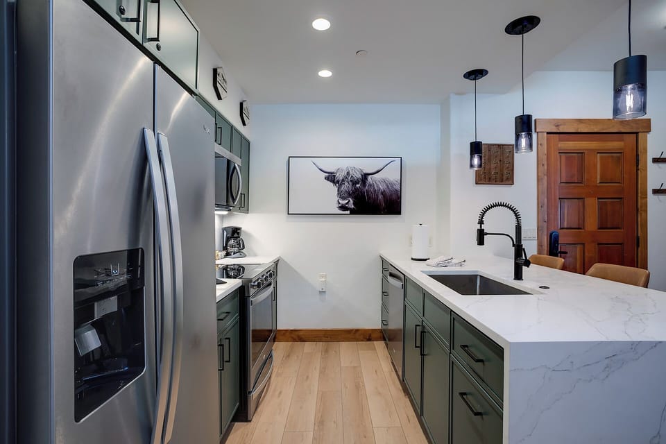 Your fully equipped kitchen blends modern style with warm wood accents, featuring stainless steel appliances, sleek green cabinetry, and a spacious marble-topped island—perfect for preparing meals or gathering with loved ones.