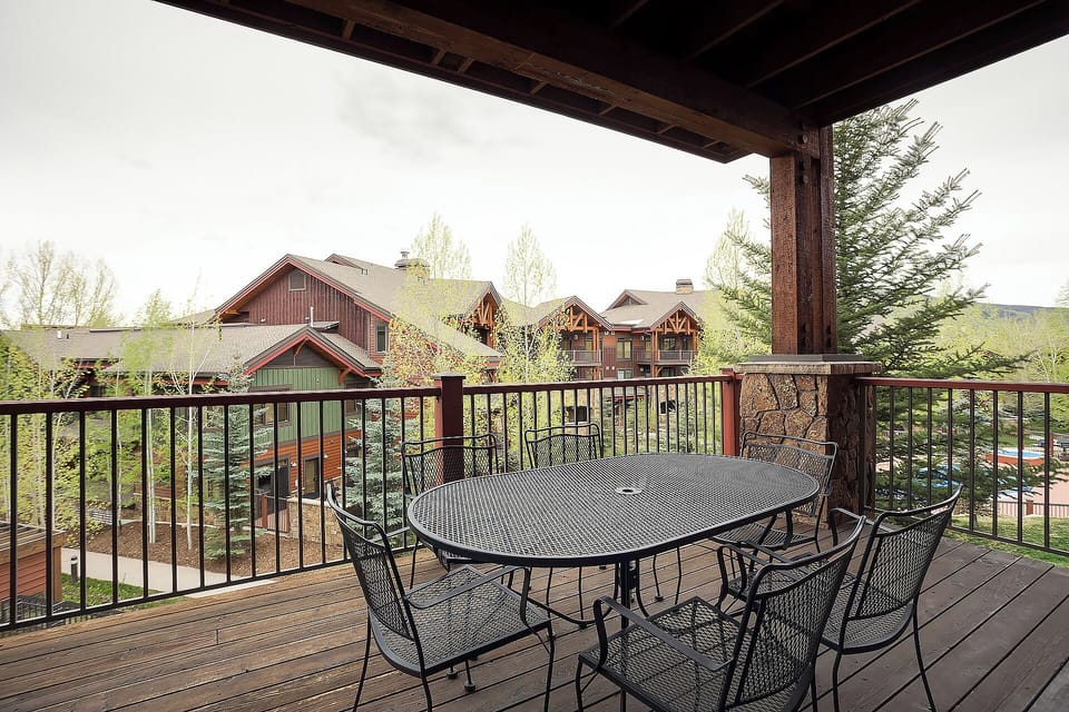 Covered deck with dining for six overlooks alpine-style architecture and mountain scenery. Spring foliage frames this peaceful outdoor space perfect for morning coffee or evening meals.