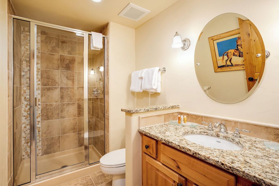 Your private bathroom features a spacious walk-in shower with natural stone tile, a granite vanity with ample counter space, and warm wood cabinetry. Fresh towels await your arrival.