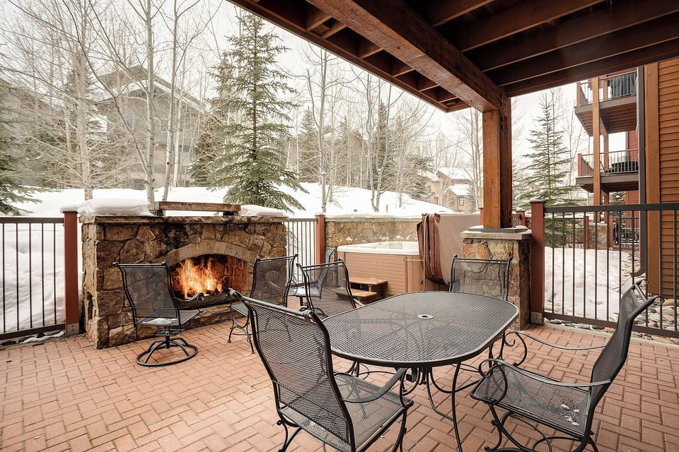 A snowy alpine setting surrounds this covered terrace with a glowing outdoor fireplace and hot tub—perfect for après-ski relaxation beneath towering evergreens and winter birches.