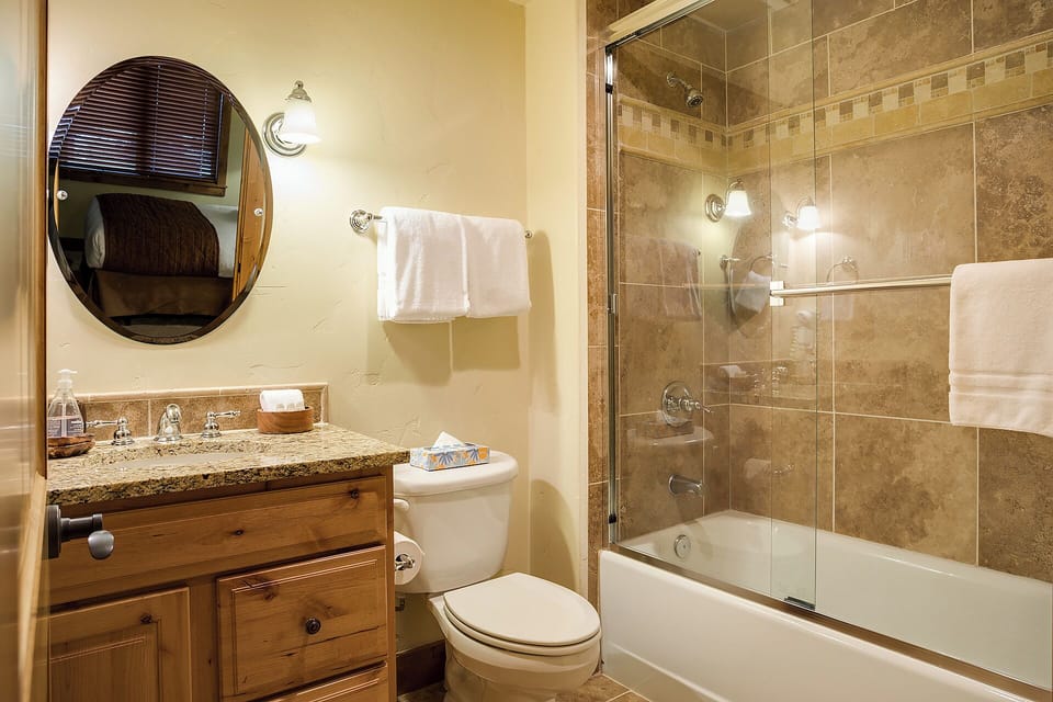 Freshen up in your private bathroom featuring natural stone tile, a tub-shower combo, and warm wood accents that create a cozy, spa-like retreat after a day of adventure.