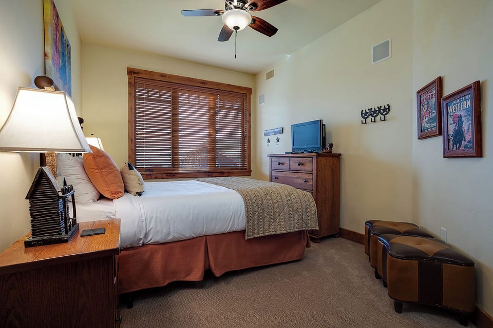 Your cozy bedroom features a plush bed with inviting linens, rustic wooden blinds filtering soft light, and Western-themed art adding character. A ceiling fan and TV offer comfort and convenience.