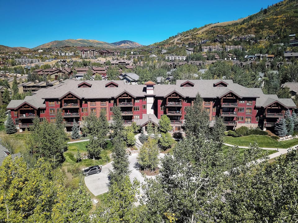 Mountain resort building nestled in a scenic alpine valley, surrounded by landscaped gardens and framed by rolling hillsides dotted with homes and natural beauty.