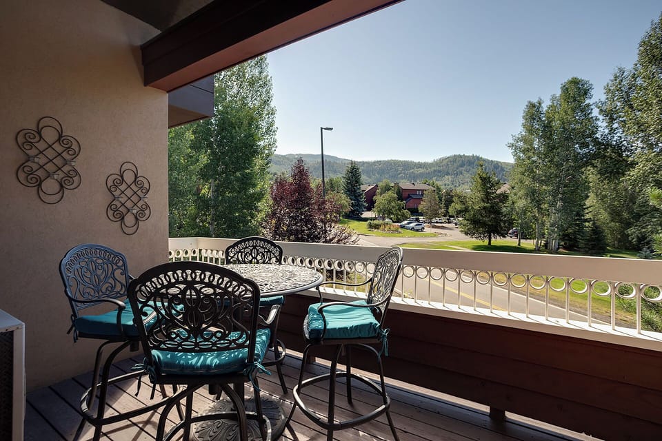 A private balcony featuring ornate wrought-iron seating with turquoise cushions overlooks a peaceful mountain neighborhood surrounded by tall trees and scenic peaks—a tranquil outdoor space for morning coffee or evening relaxation.