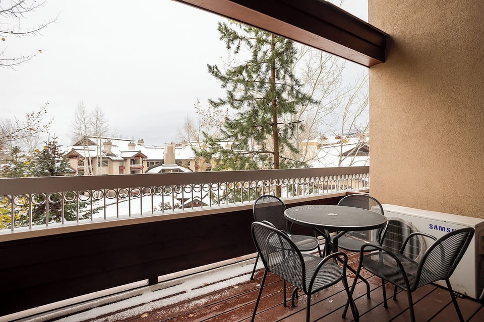 A covered balcony featuring outdoor furniture and winter views of neighboring alpine-style buildings and snow-dusted pine trees beyond the railing.