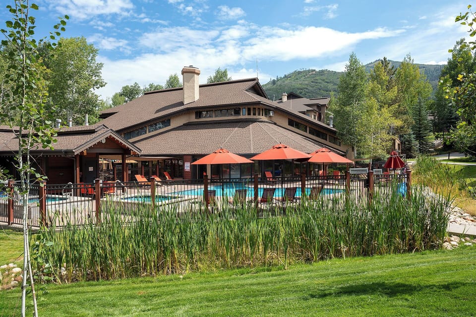 A mountain resort with an outdoor pool surrounded by orange umbrellas and lounge seating, set against a backdrop of forested peaks and aspens.