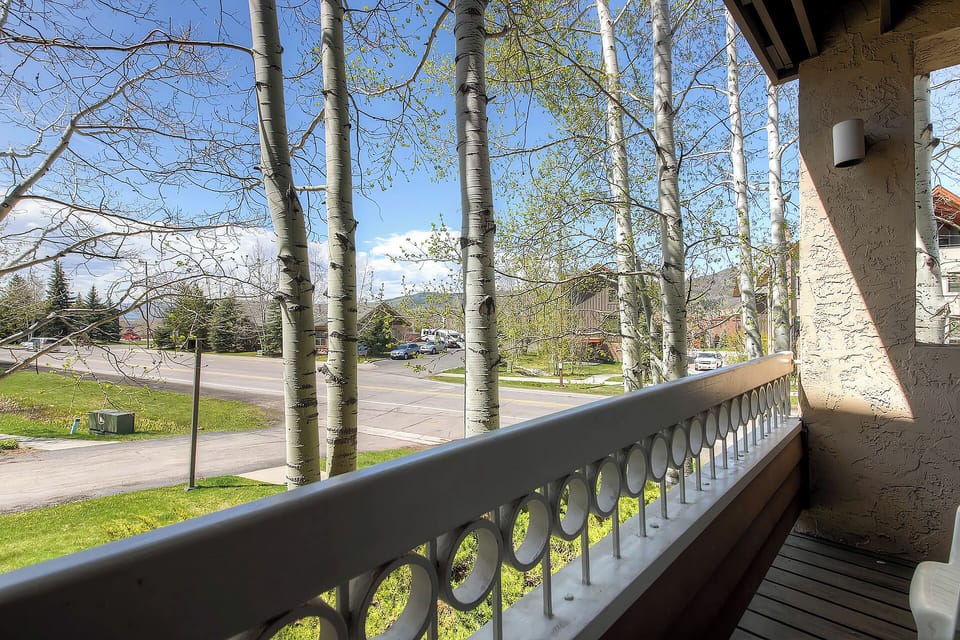 This balcony overlooks a quiet residential street framed by towering aspens and evergreens. Bright spring foliage and mountain peaks in the distance complete the peaceful neighborhood setting.