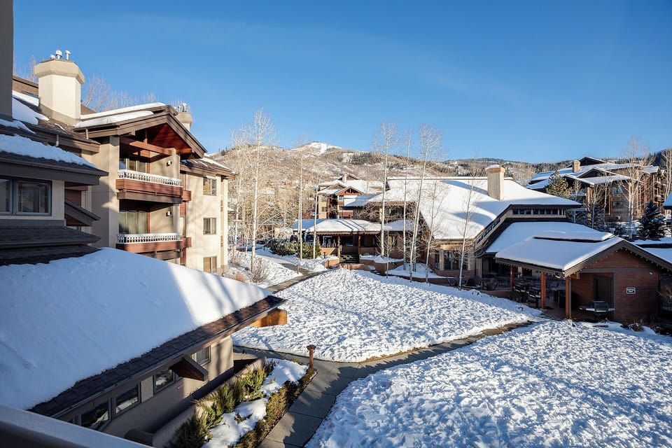 A pristine winter scene showcasing the property nestled among snow-covered Alpine-style homes, with frosted trees and ski slopes rising in the background under a clear blue sky.