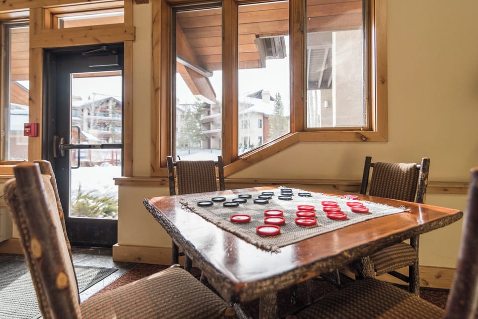 The communal game room overlooks a snowy alpine village, offering rustic wood finishes and a checkers table where you can relax with fellow travelers while enjoying mountain views through expansive windows.