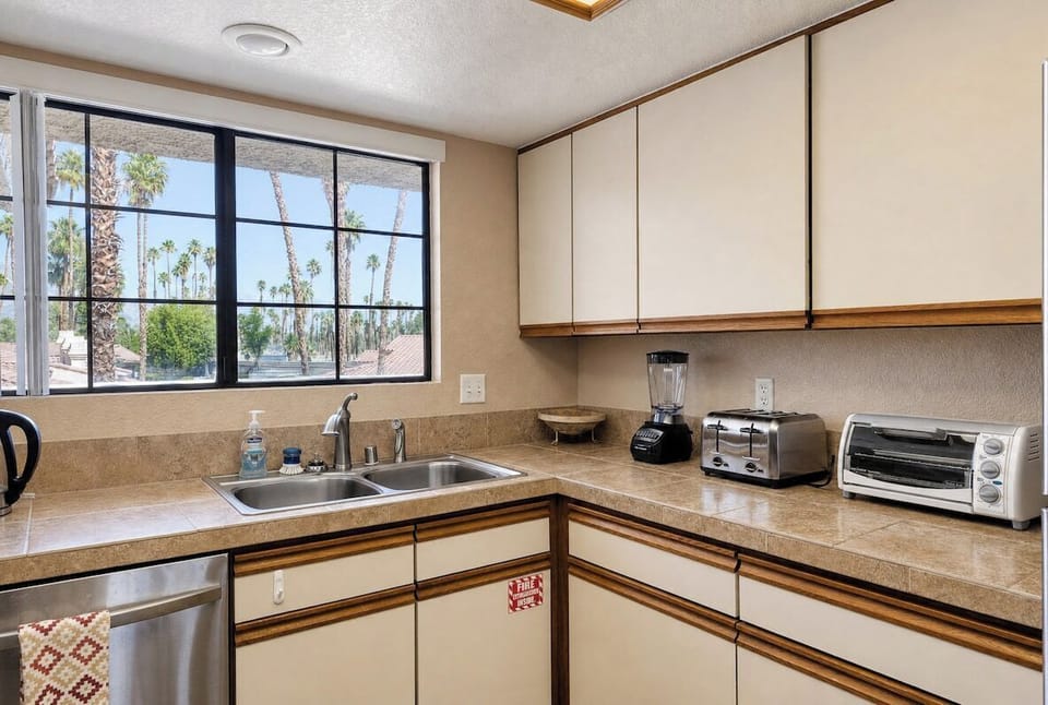 Your fully equipped kitchen features modern appliances and ample counter space for preparing meals, with tropical palm views through large windows that fill the space with natural light.