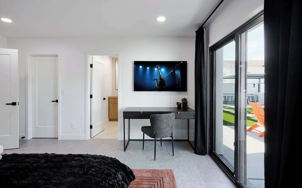 Primary bedroom with King bed, designer furnishings, walk-in closet, smart TV, ceiling fan, direct access to backyard, work station, and en-suite bathroom. 