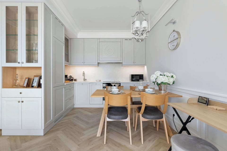 A modern kitchenette and dining area with white cabinets, a wooden dining table, and several chairs. The open layout connects the kitchen with the bright living space.