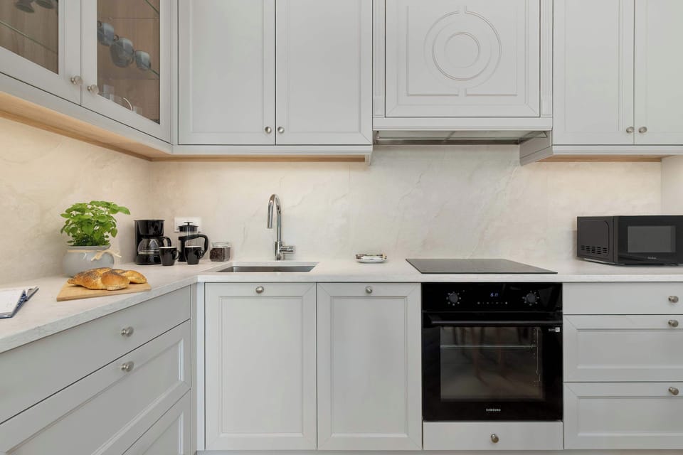 A bright white kitchenette with modern cabinets, a built-in oven, microwave, and countertop appliances. Light wood flooring and small decorative plants add warmth to the space.