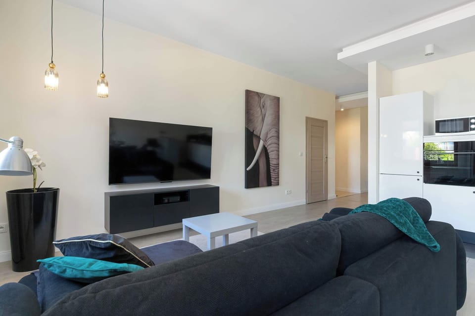 A modern living room with a TV, comfortable sofa, and dining area. The large windows fill the space with natural light.