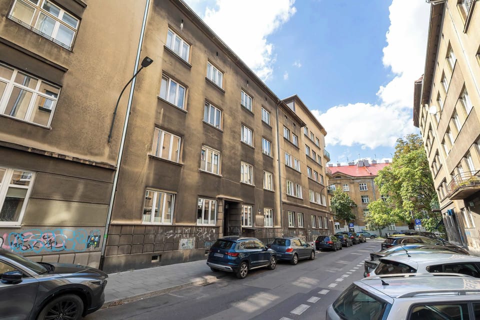 An exterior shot displays the street view of the apartment building with a classic brown facade. Cars are parked along the road beneath a clear blue sky, showing the urban neighborhood.