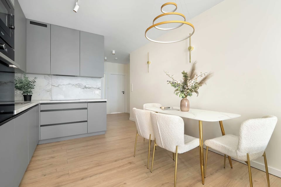 A bright dining area features a round white table surrounded by four plush, tufted chairs, situated next to the kitchen. A golden ring chandelier hangs elegantly overhead.