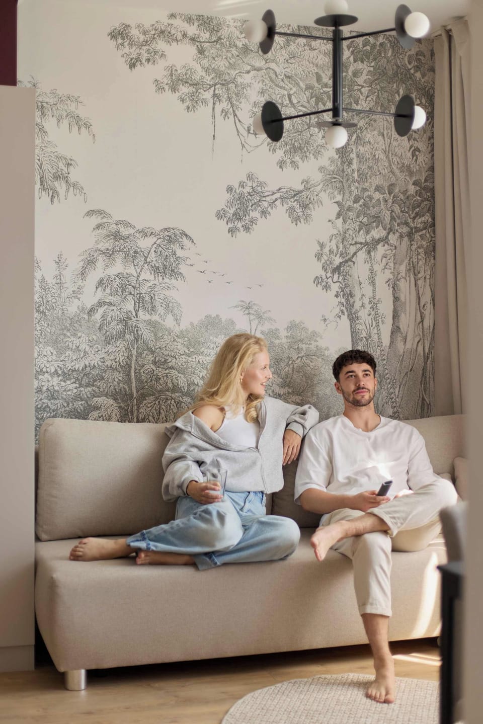 Stylowy i komfortowy salon z wygodną sofą, dużymi oknami i elegancką strefą wypoczynku. Ciepła drewniana podłoga, dekoracyjna tapeta, burgundowe akcenty i nastrojowe oświetlenie nadają wnętrzu charakter.
