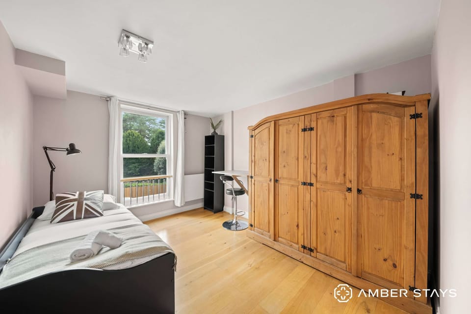 This bright bedroom features a single bed, a large freestanding wooden wardrobe, a tall shelving unit, and a window overlooking greenery.