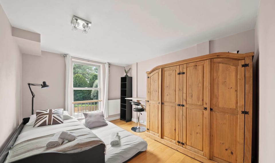 This bright bedroom features a versatile trundle bed setup with neutral bedding and a Union Jack pillow, situated opposite an expansive, classic wooden wardrobe.