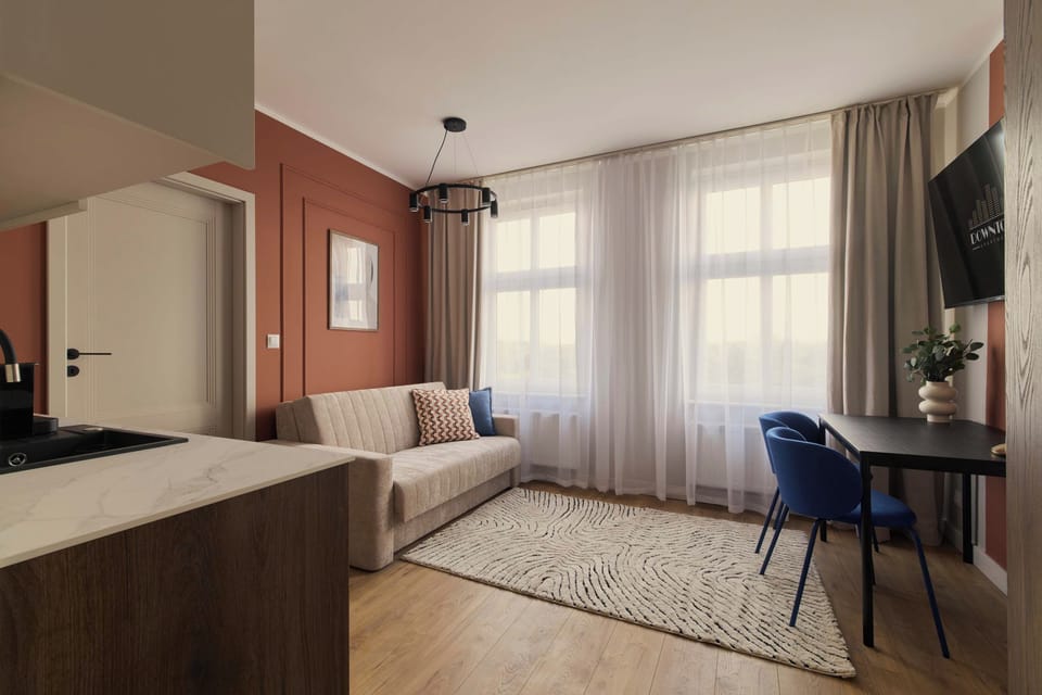 Stylowy i przytulny salon połączony z aneksem kuchennym, utrzymany w ciepłej kolorystyce. Drewniane akcenty, dekoracyjna ceglana ściana, wygodna sofa i duże okno tworzą jasną, komfortową przestrzeń do wypoczynku.